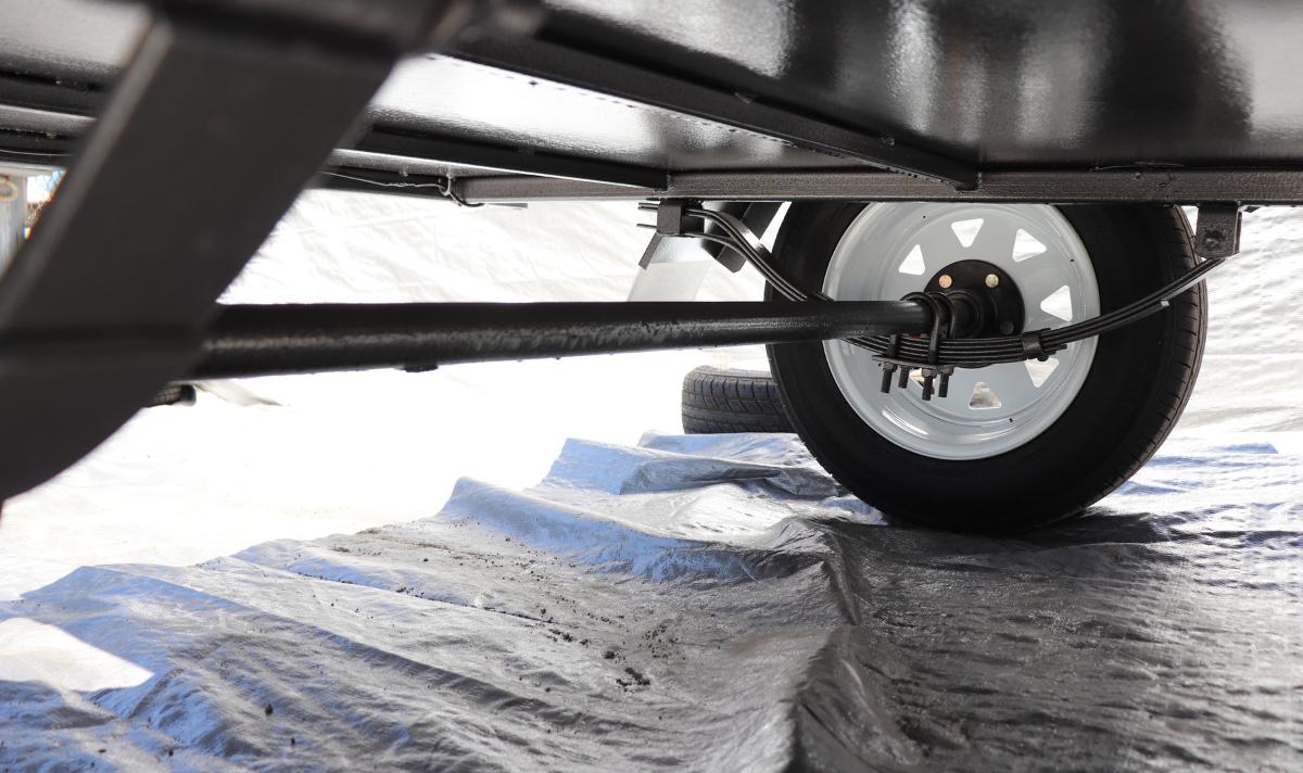 Underside view of a trailer showcasing the suspension system, axle, and wheel assembly on a black tarpaulin background.