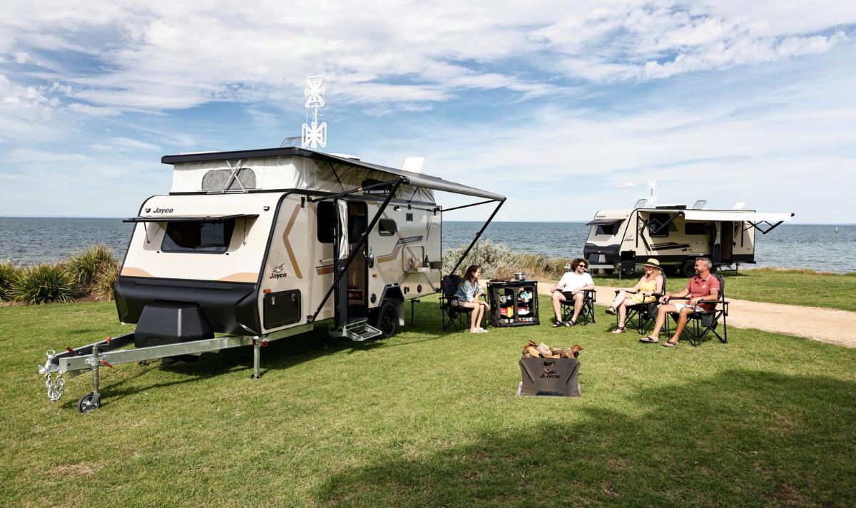Camping scene featuring two Jayco caravans parked by the water, with four people relaxing in chairs. A portable fire pit sits nearby, surrounded by lush green grass and a scenic view of the lake under a partly cloudy sky.